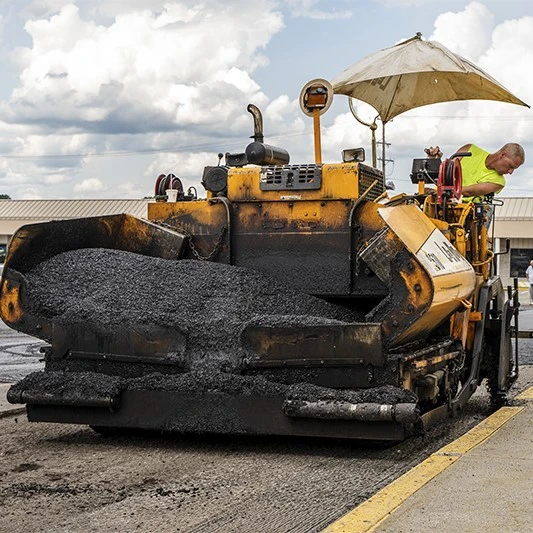 Worker operating heavy machinery for asphalt repair and paving under a cloudy sky, improving a parking lot surface.